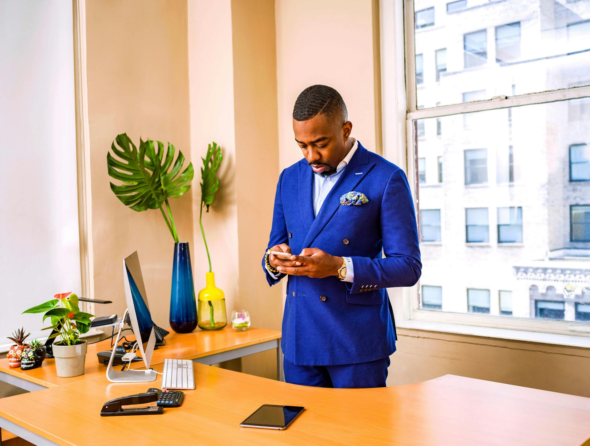 Businessman in a blue suit using a smartphone at his office desk with city view.