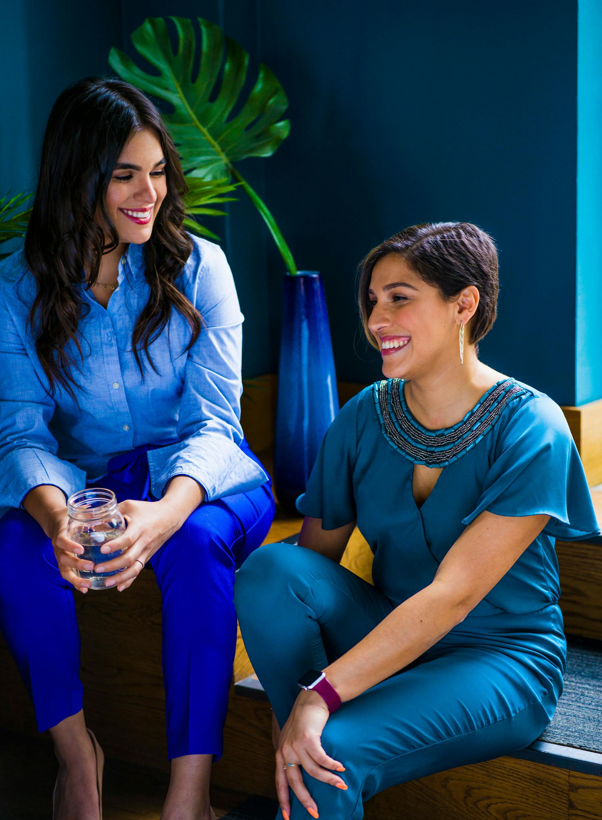 Two women sitting and smiling while having a conversation in a stylish, modern office space, reflecting a relaxed and engaging work environment.