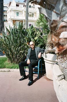 阳光明媚，一位戴着太阳镜的成年男子坐在户外，背景是棕榈树和建筑物