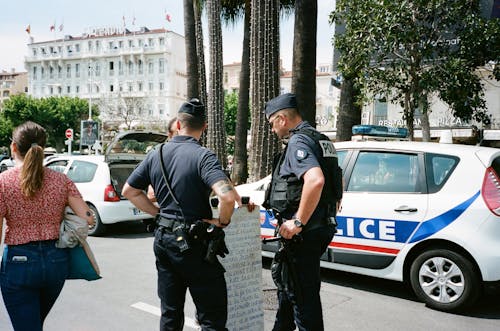 двое полицейских стоят рядом с другим человеком рядом с бело синей полицейской машиной на дороге