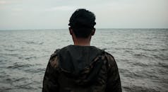 一名穿着迷彩夹克的男子站在海滩上，望着海天相接的地平线