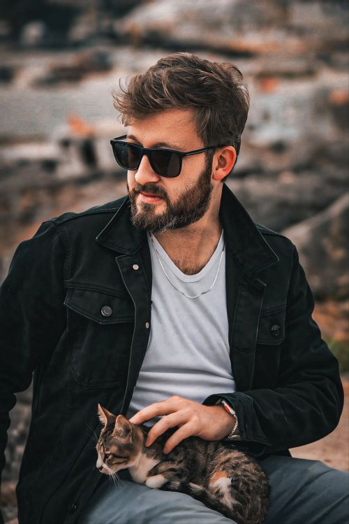Free A fashionable man with sunglasses sits petting a cat in an urban environment. Stock Photo