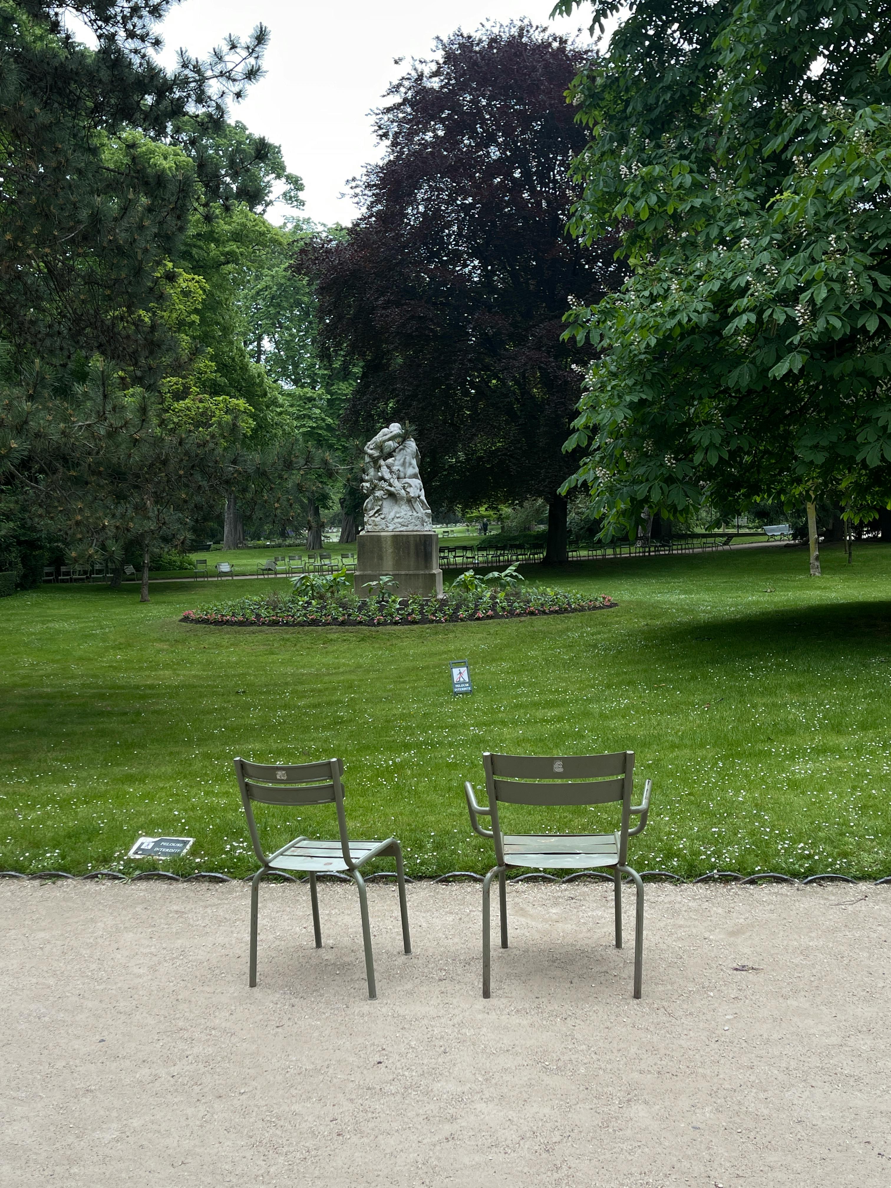 A tranquil park scene with two chairs facing a statue surrounded by lush greenery.