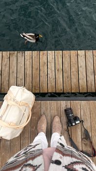 Top-down view of a person with camera, bag, and duck on a pier in Serbia. Captures tranquil outdoor scene.