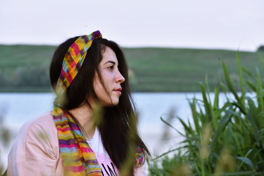 Side View Photo of Woman in Pink Top and Multi-colored Headscarf Sitting Next to Tall Grass With Body of Water in the Background