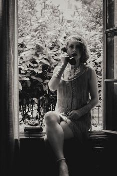 Elegant woman in vintage dress sitting by window using retro telephone.