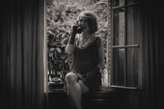 A woman in flapper attire sits by a window using a vintage telephone.