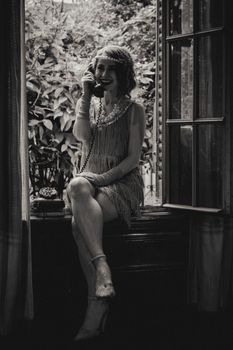 Black and white photo of a smiling woman in vintage attire posing with a retro telephone by a window.