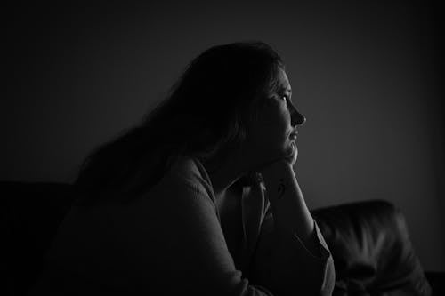 Grayscale Photo of Woman Sitting on Sofa With Her Head On Her Hand Looking Away