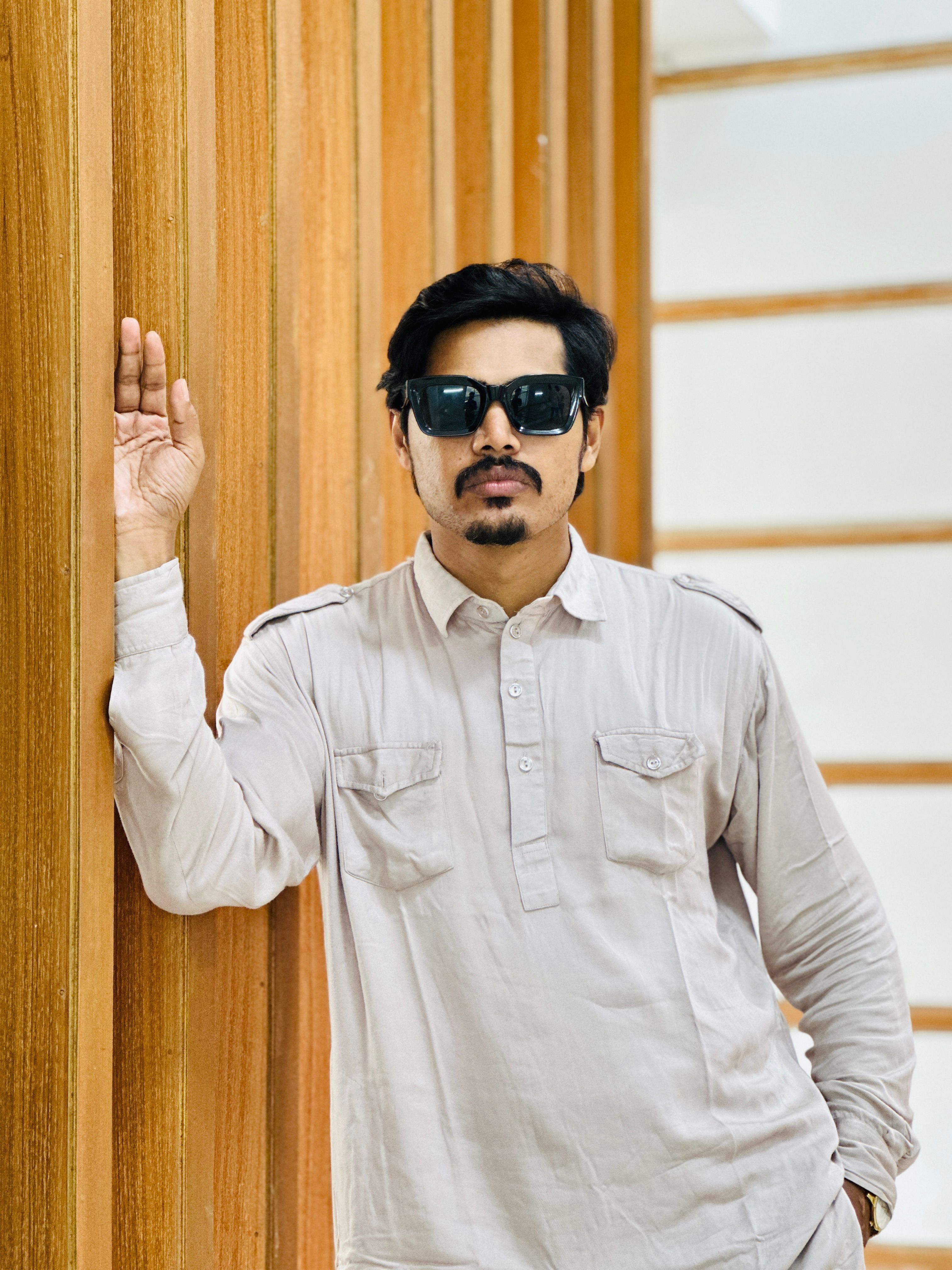 Portrait of a stylish man with sunglasses posing against a wooden wall indoors.
