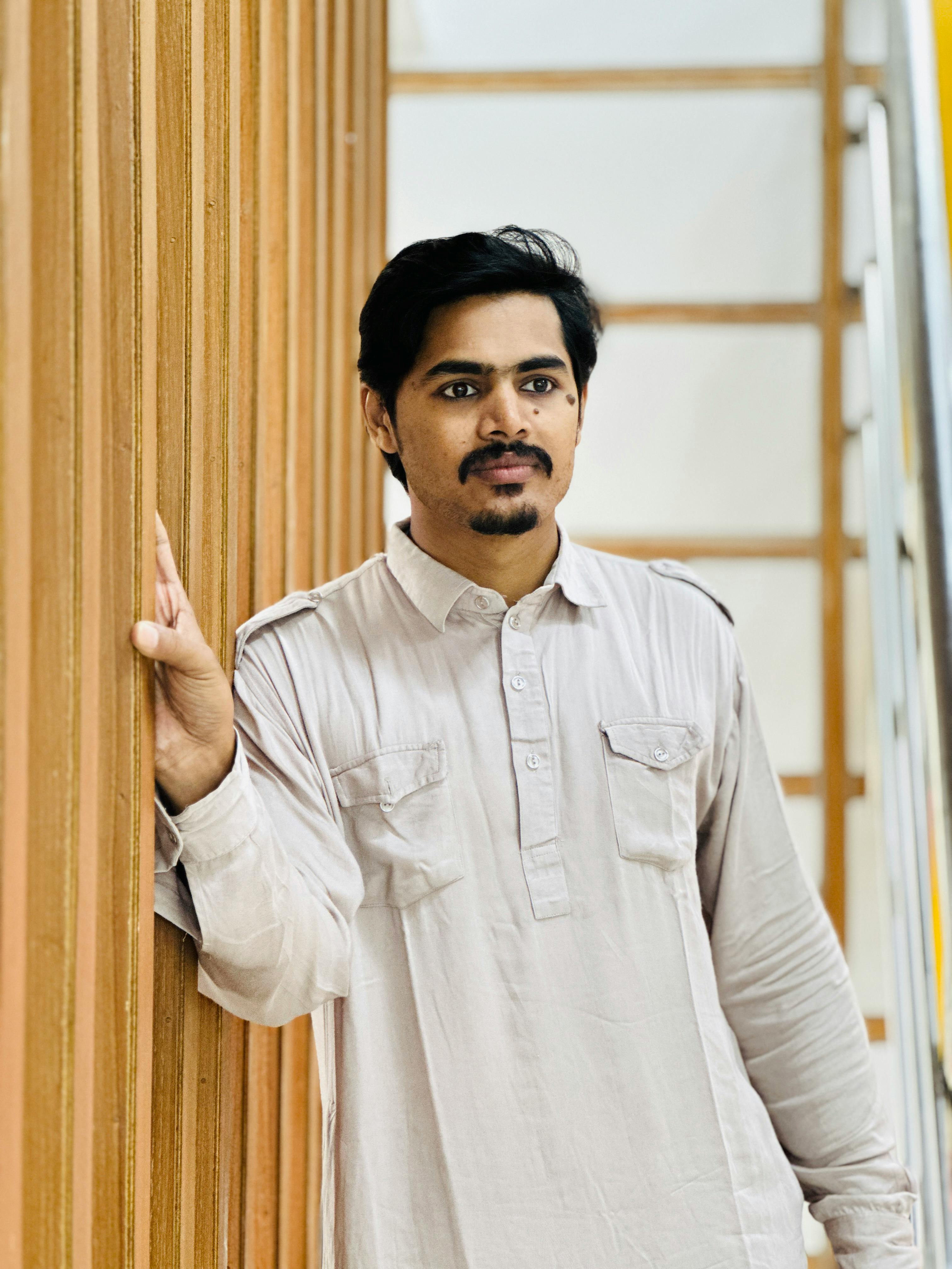 A thoughtful man in traditional South Asian attire stands against a wooden wall indoors.