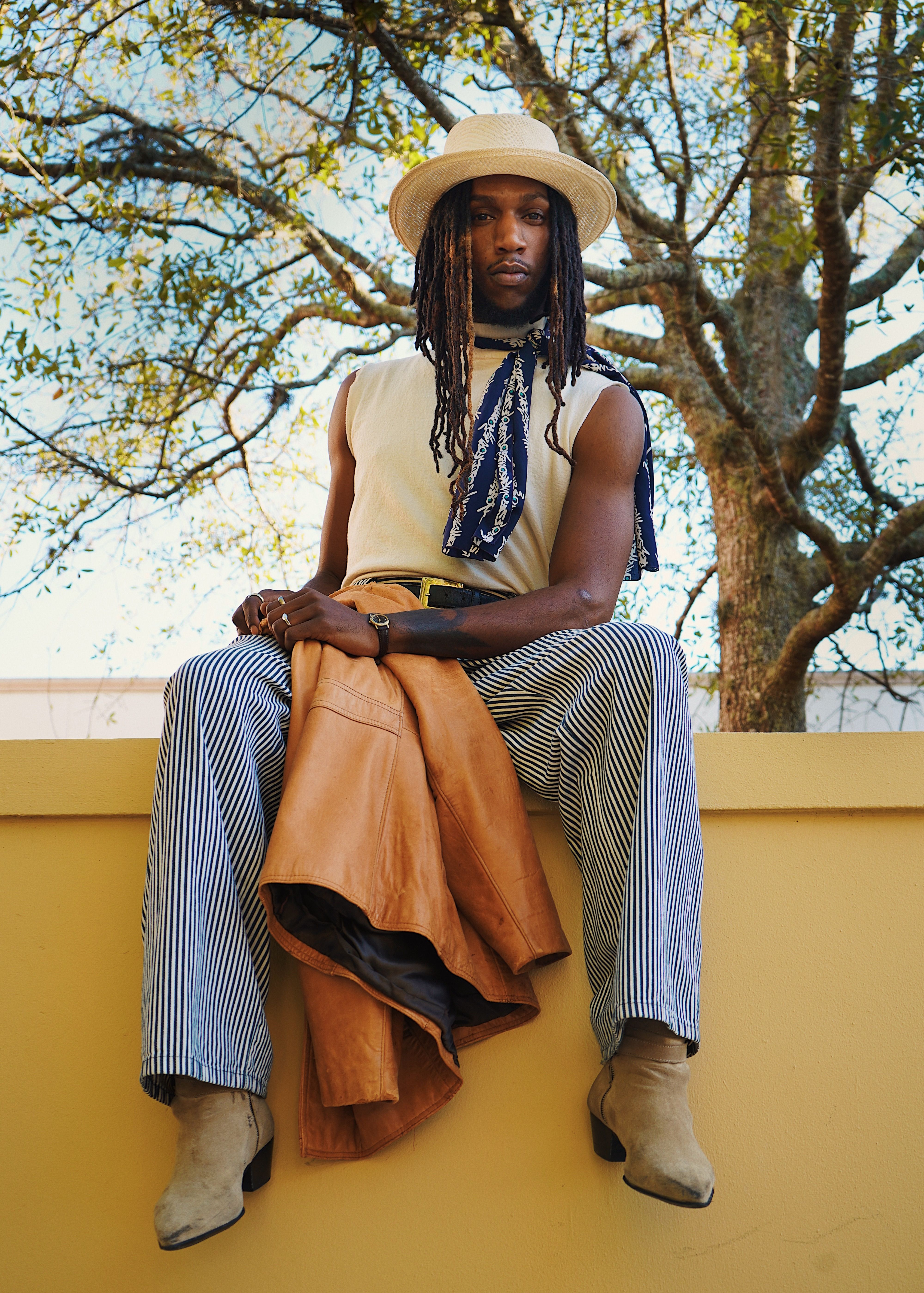 A fashionable man in urban attire sits on a wall with trees in the background.