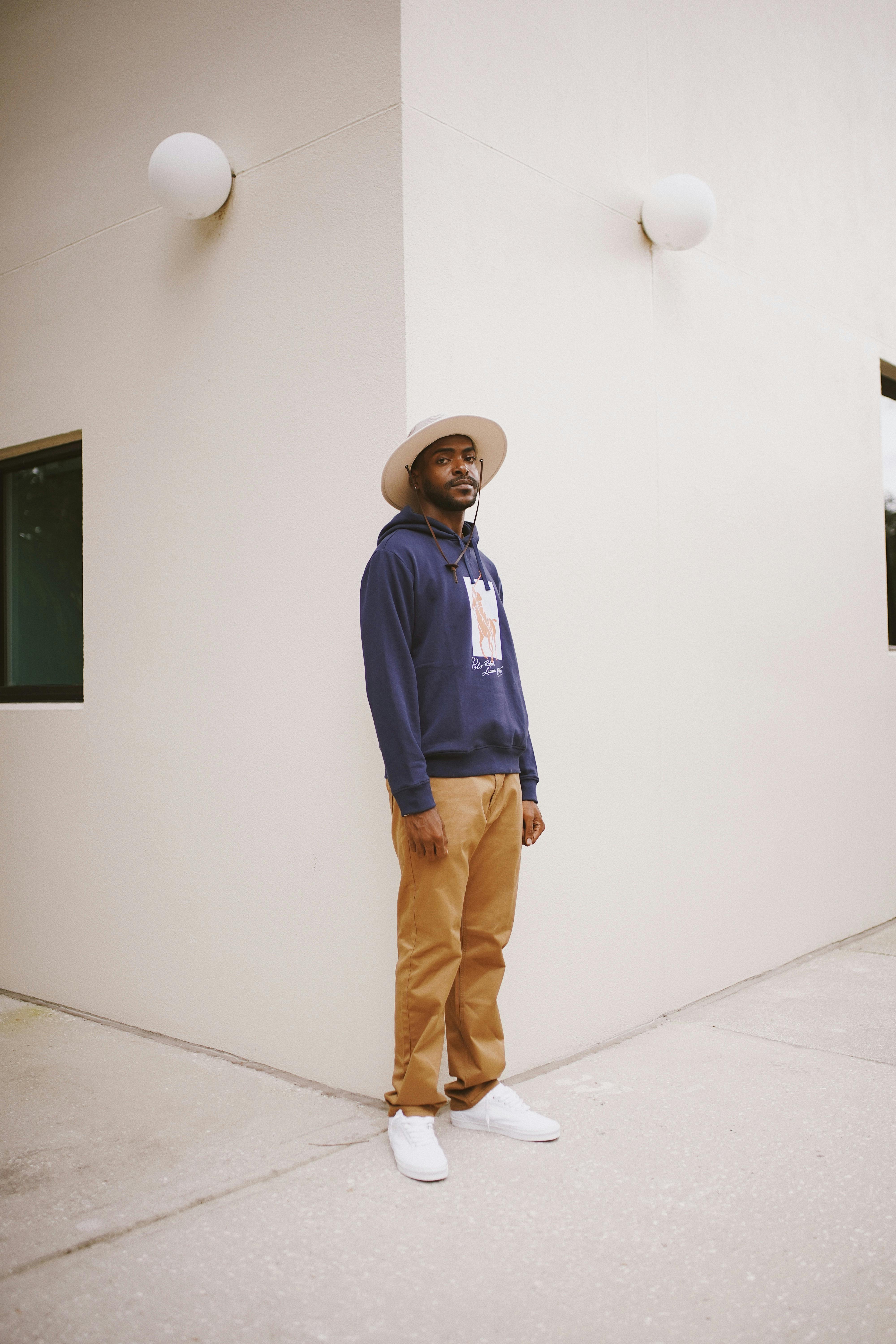 Stylish man wearing hat and hoodie posing outdoors against modern architecture.