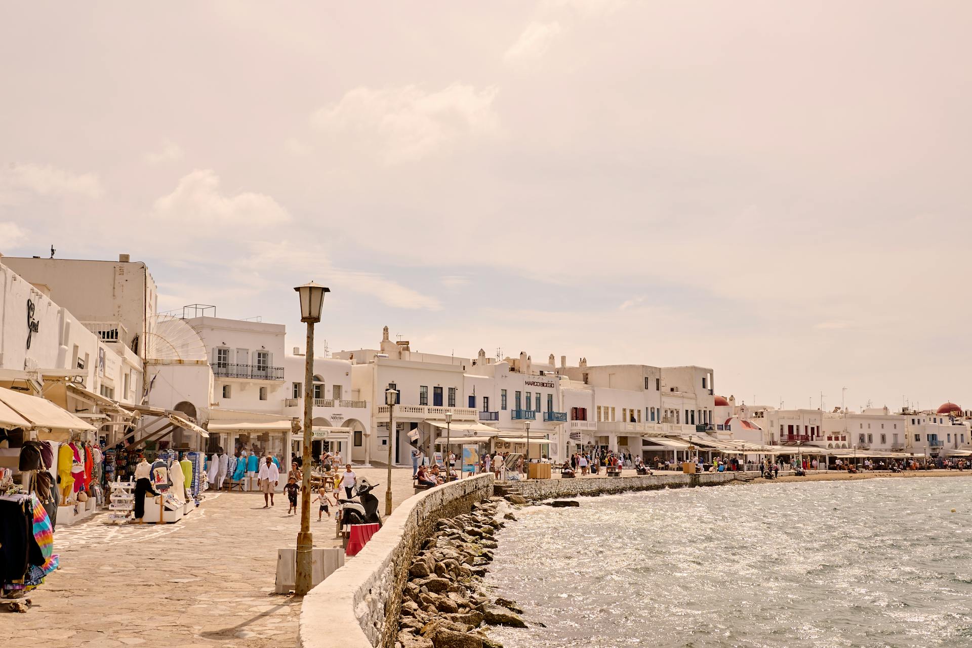 Bustling summer scene along the picturesque Mykonos waterfront promenade with shops and people.