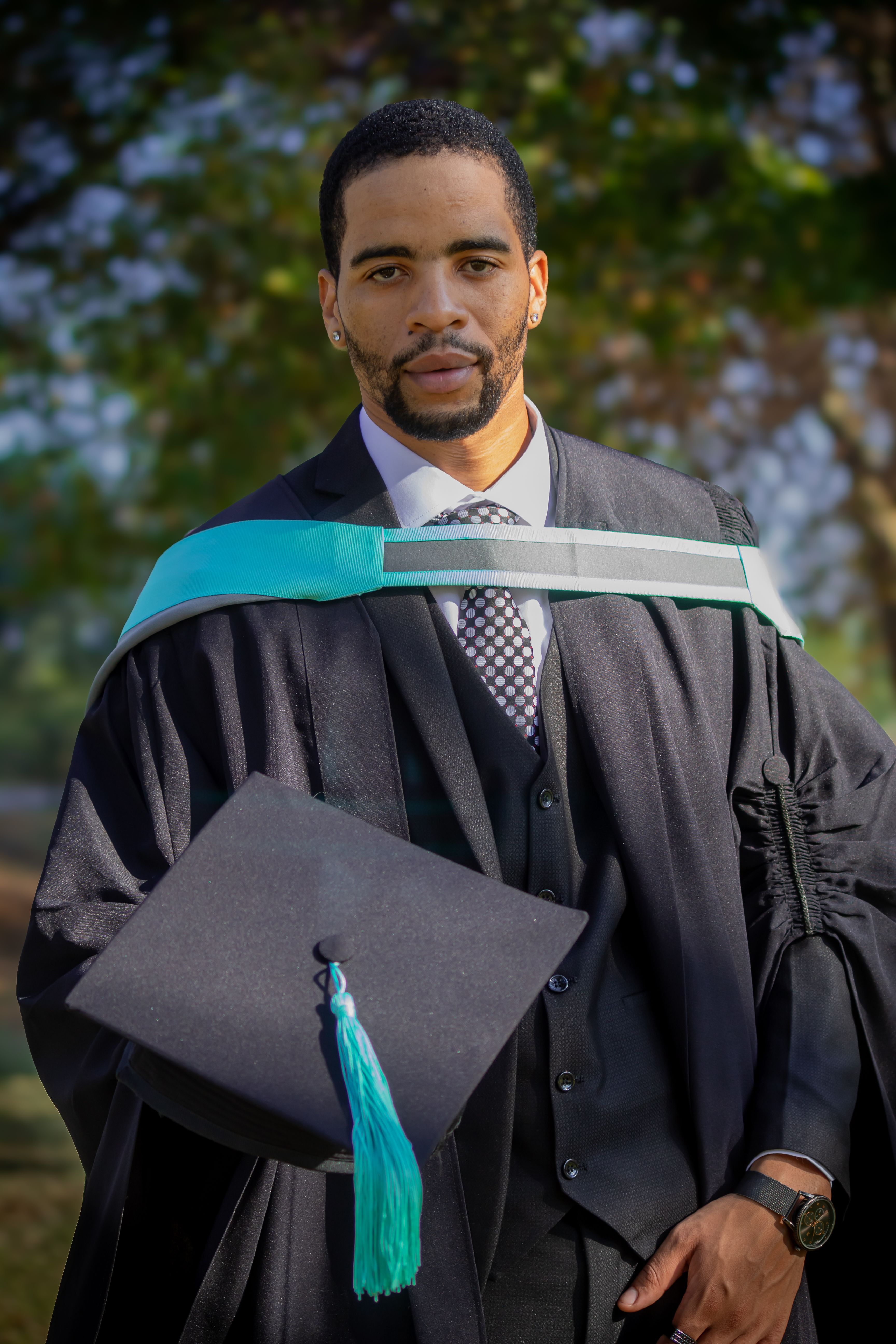 A young man in a graduation gown holding an academic cap outdoors in Pretoria, South Africa.