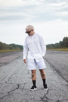 A person with blonde hair in sunglasses stands on a rural road wearing white attire.