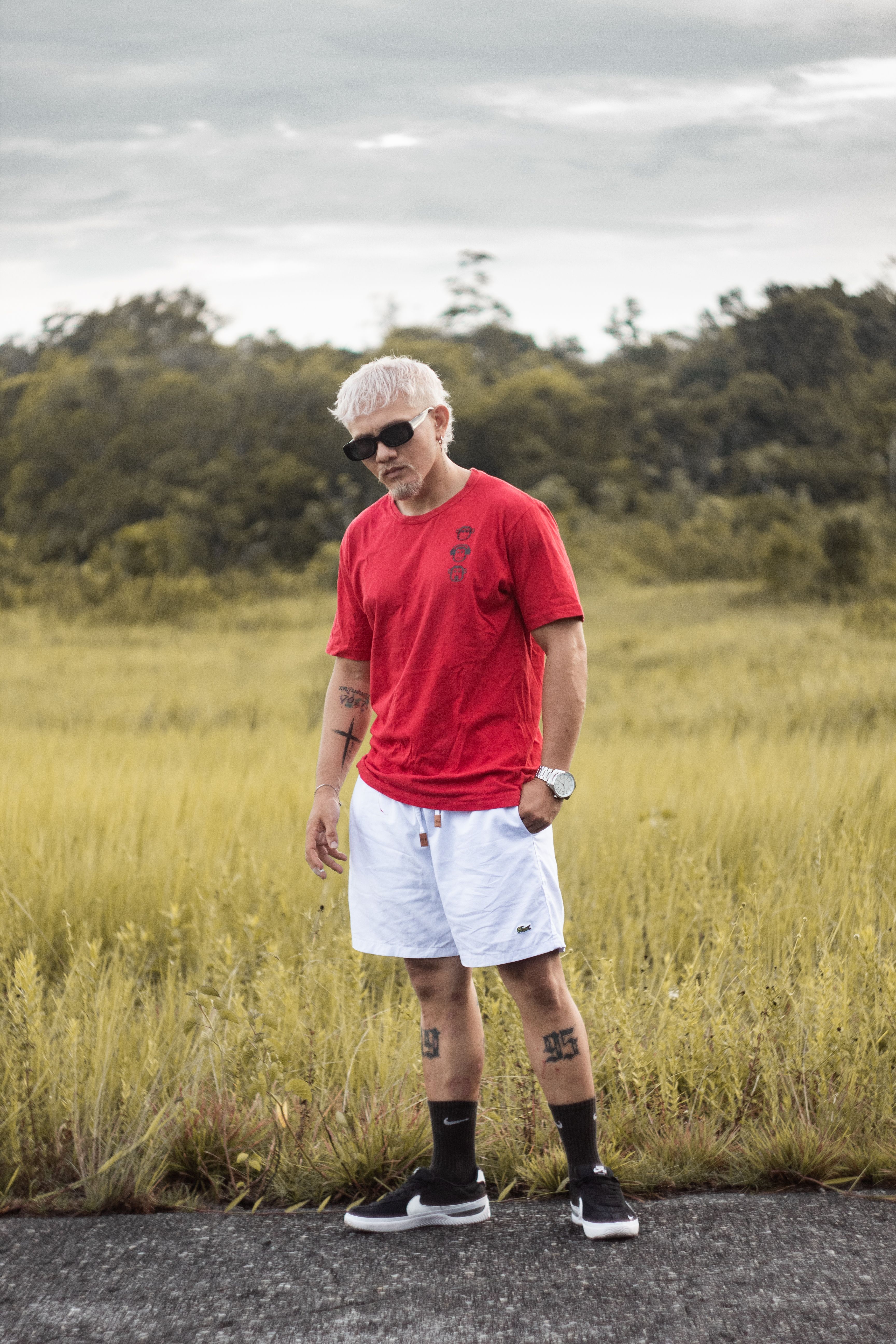 Stylishly dressed man in a red t-shirt and shorts standing outdoors in a rural field.