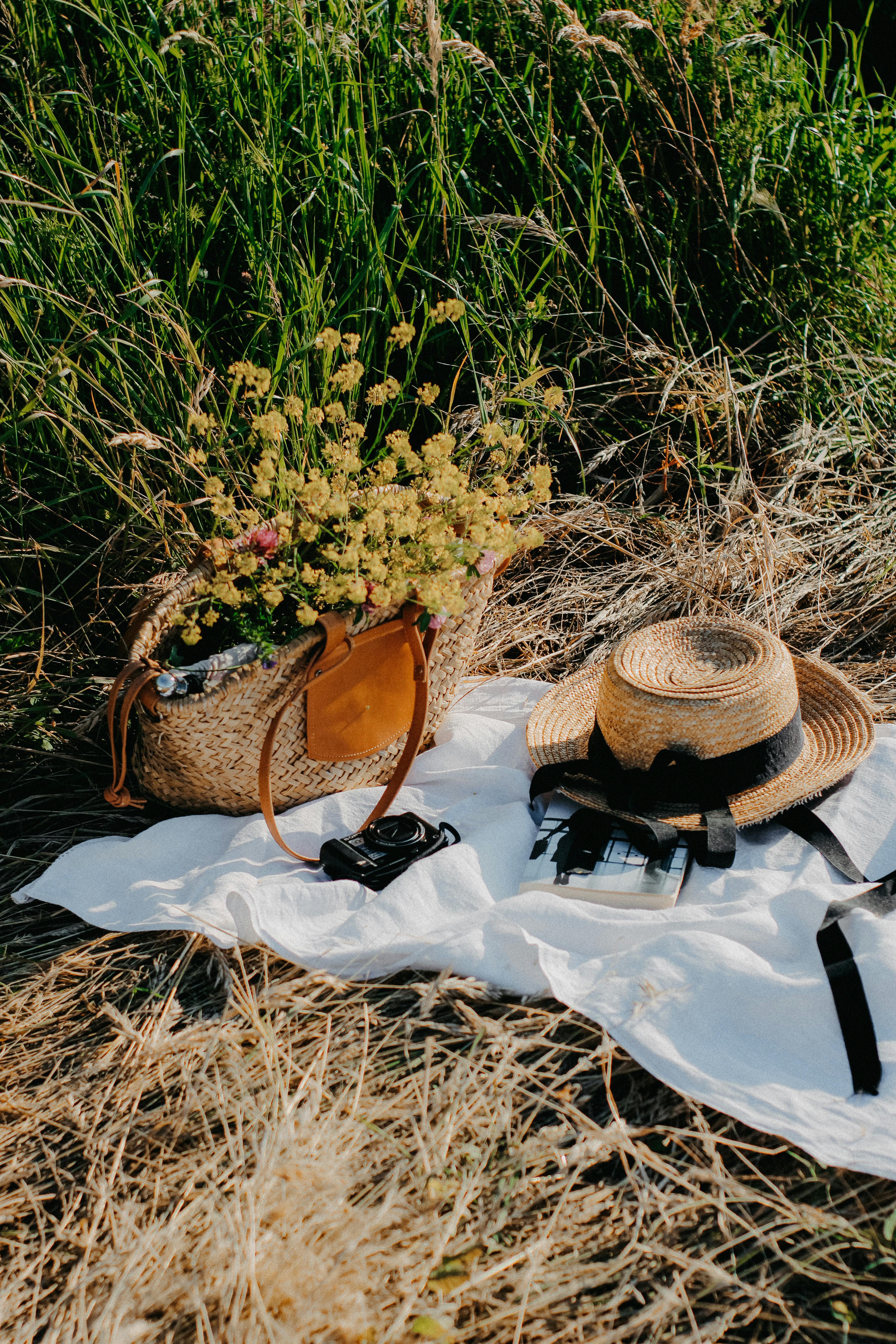 Sunlit picnic with a straw hat, basket of flowers, and camera on a blanket in a grassy field.