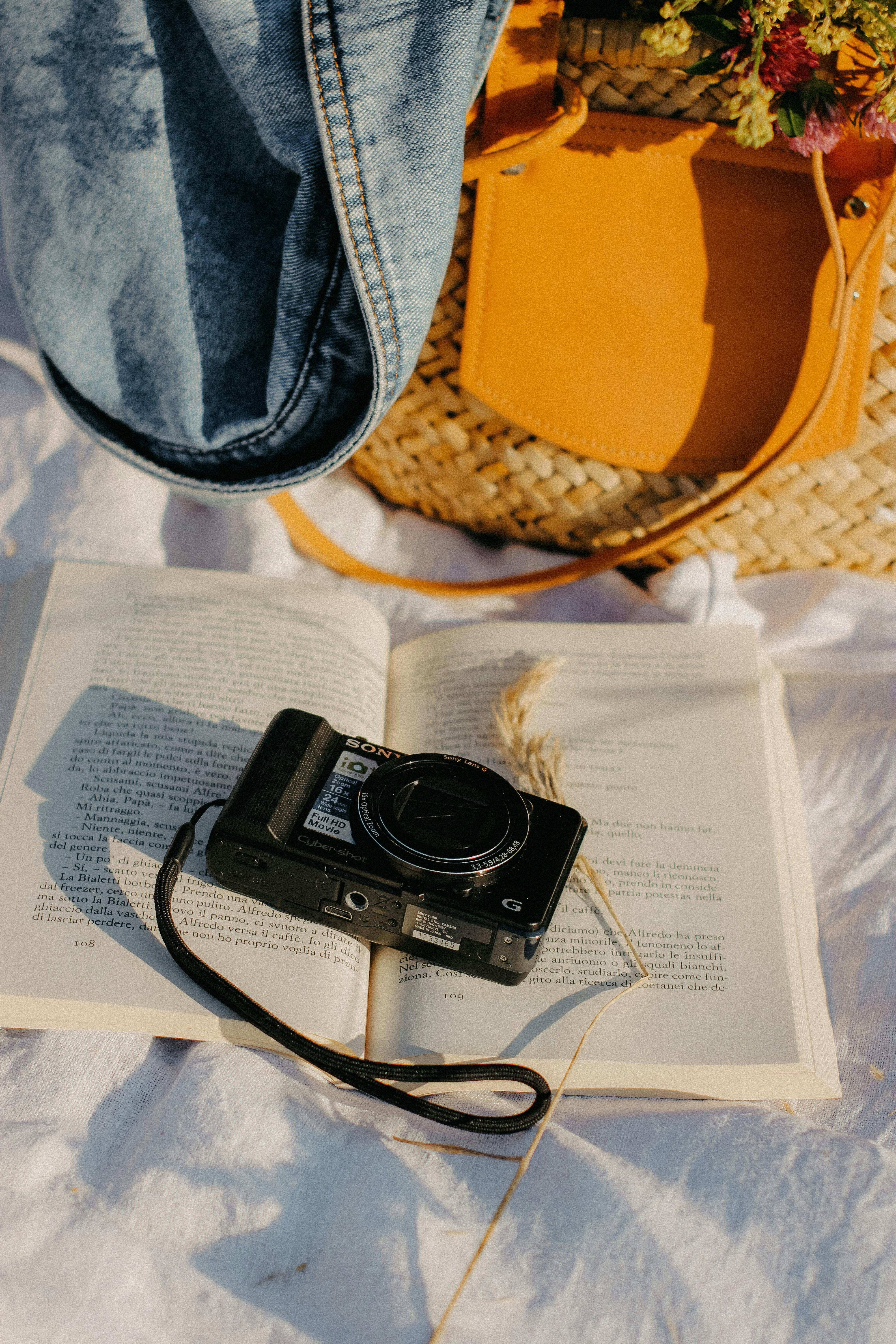 An artistic composition of a vintage camera on an open book, laid on a picnic blanket with a woven bag.
