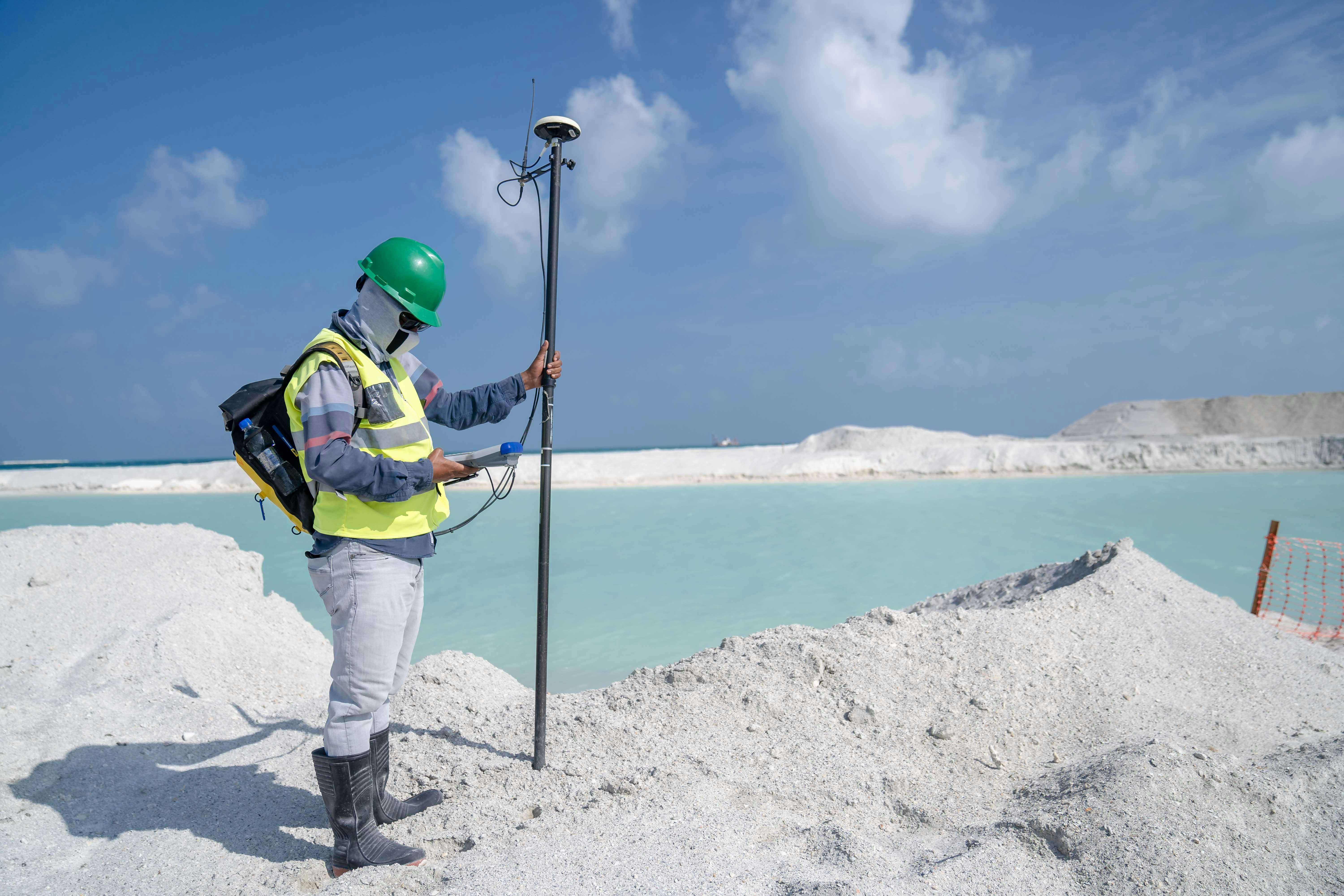 Surveyor in protective gear conducting measurements by a remote beach with GPS equipment.