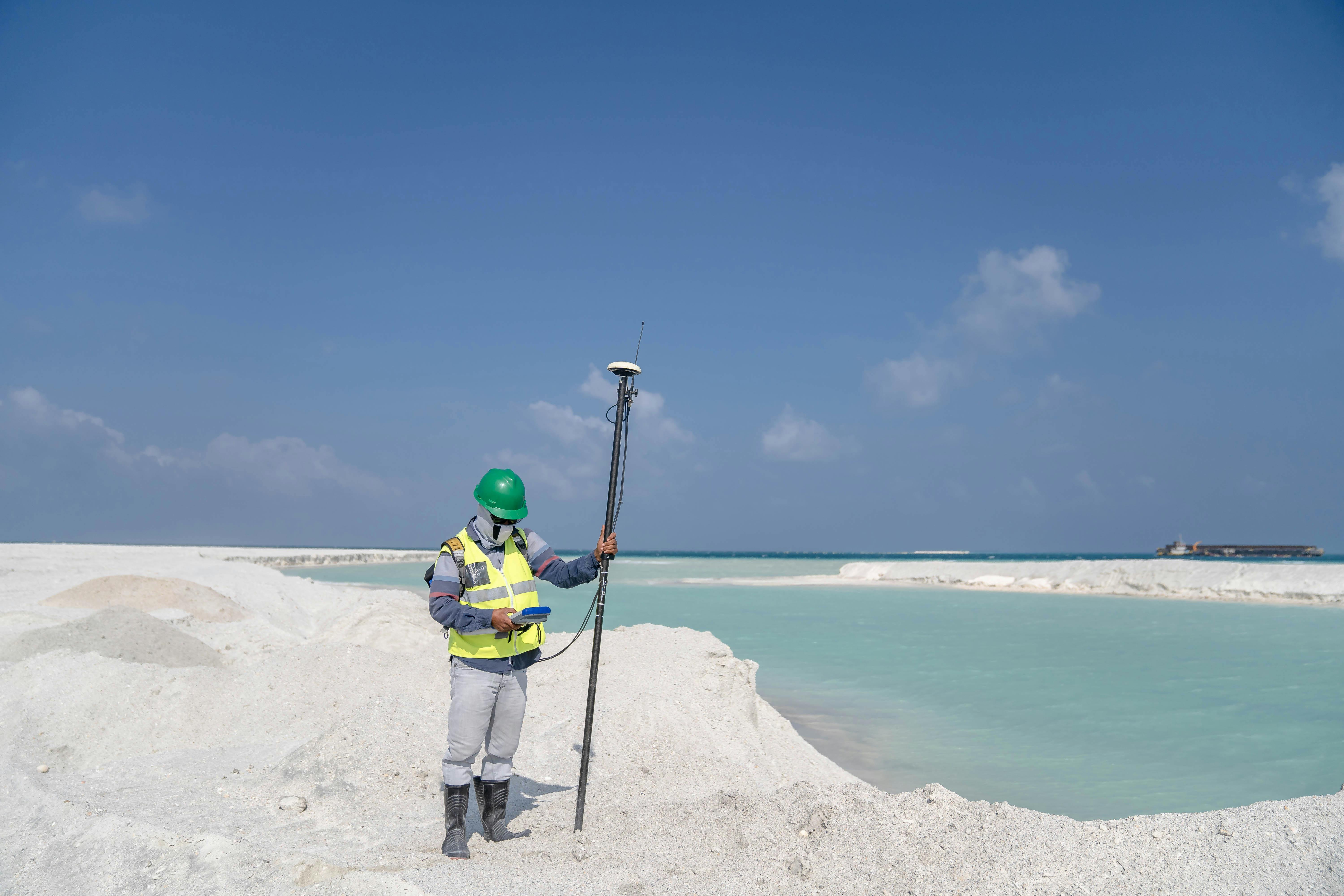 Surveyor in safety gear measuring near a turquoise lake under a clear sky.