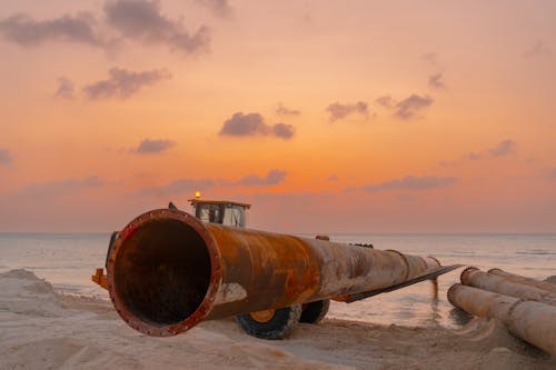 A bulldozer moving large pipes on a beach construction site with a stunning sunset backdrop.  Bulldozer moving large pipes on a beach construction site during sunset.