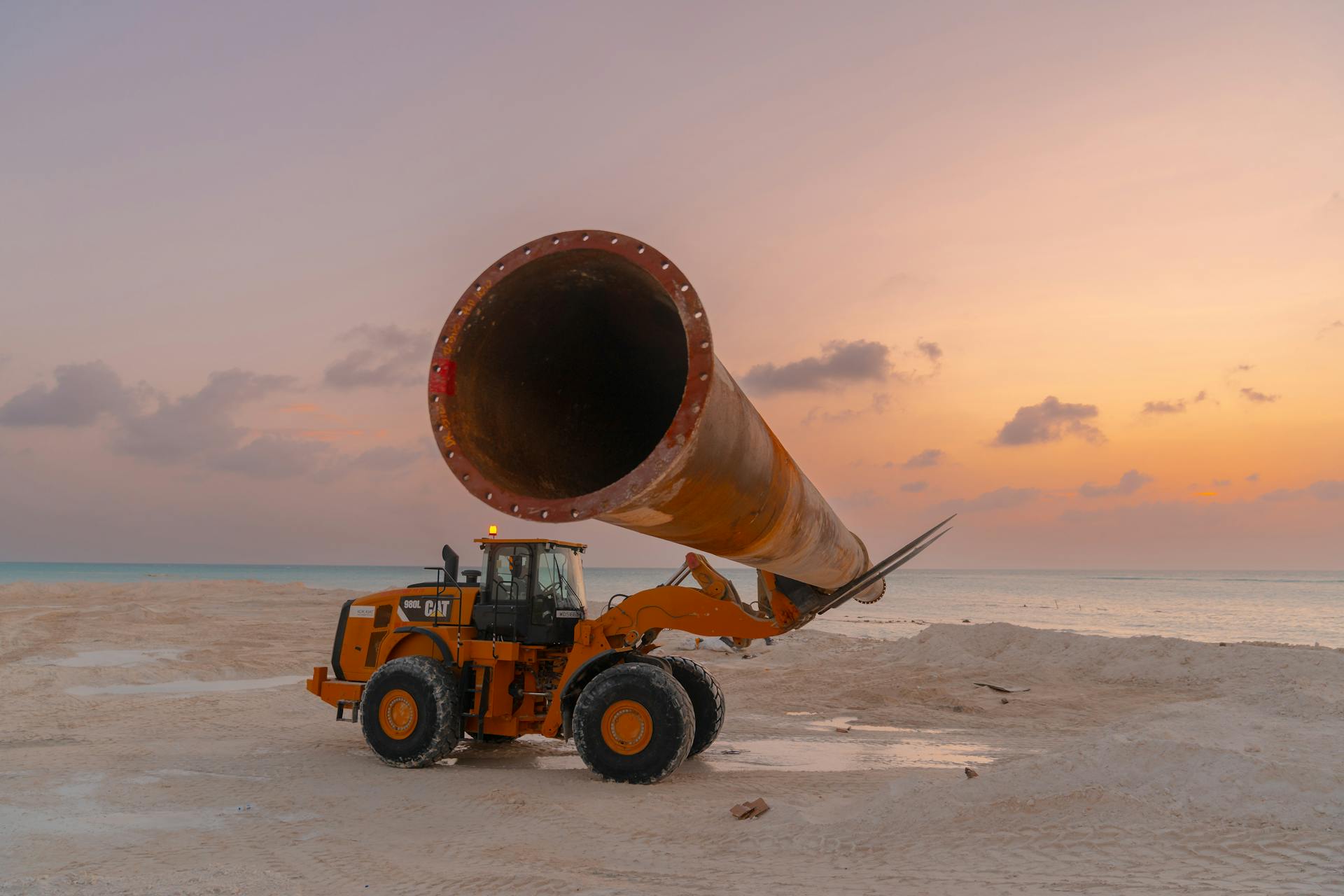 Construction equipment at sunset on a beach, featuring heavy machinery and large pipe work.