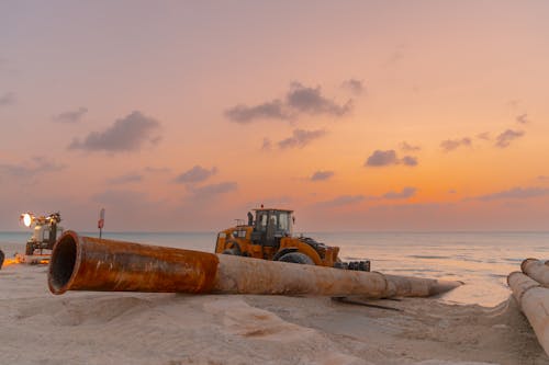 A bulldozer moving large pipes on a beach construction site with a stunning sunset backdrop.  Bulldozer moving large pipes on a beach construction site during sunset.