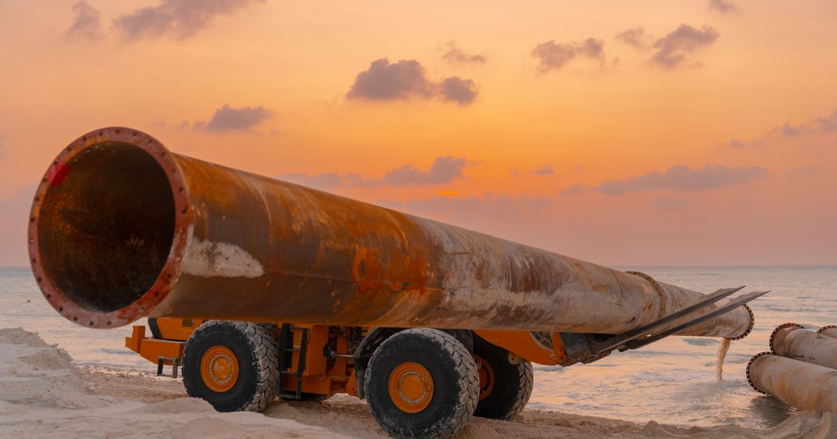 Um duto enferrujado e máquinas em um canteiro de obras costeiro durante o pôr do sol.