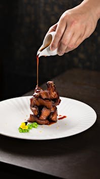 Elegant close-up of hand pouring rich sauce over a gourmet dish with roasted meat on a white plate.