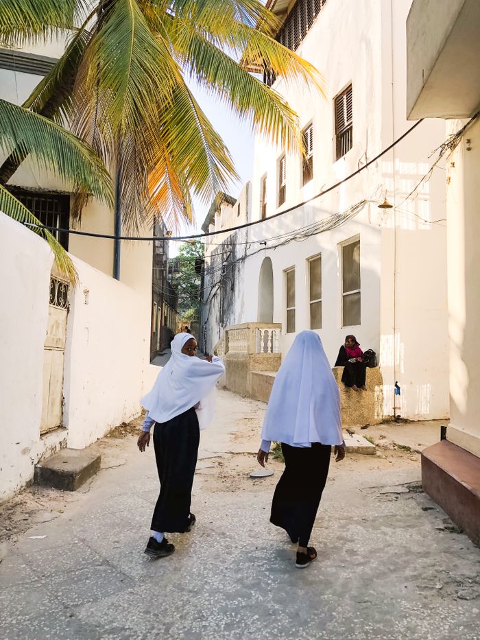 Two women in headscarves walking through a sunlit alley in Zanzibar