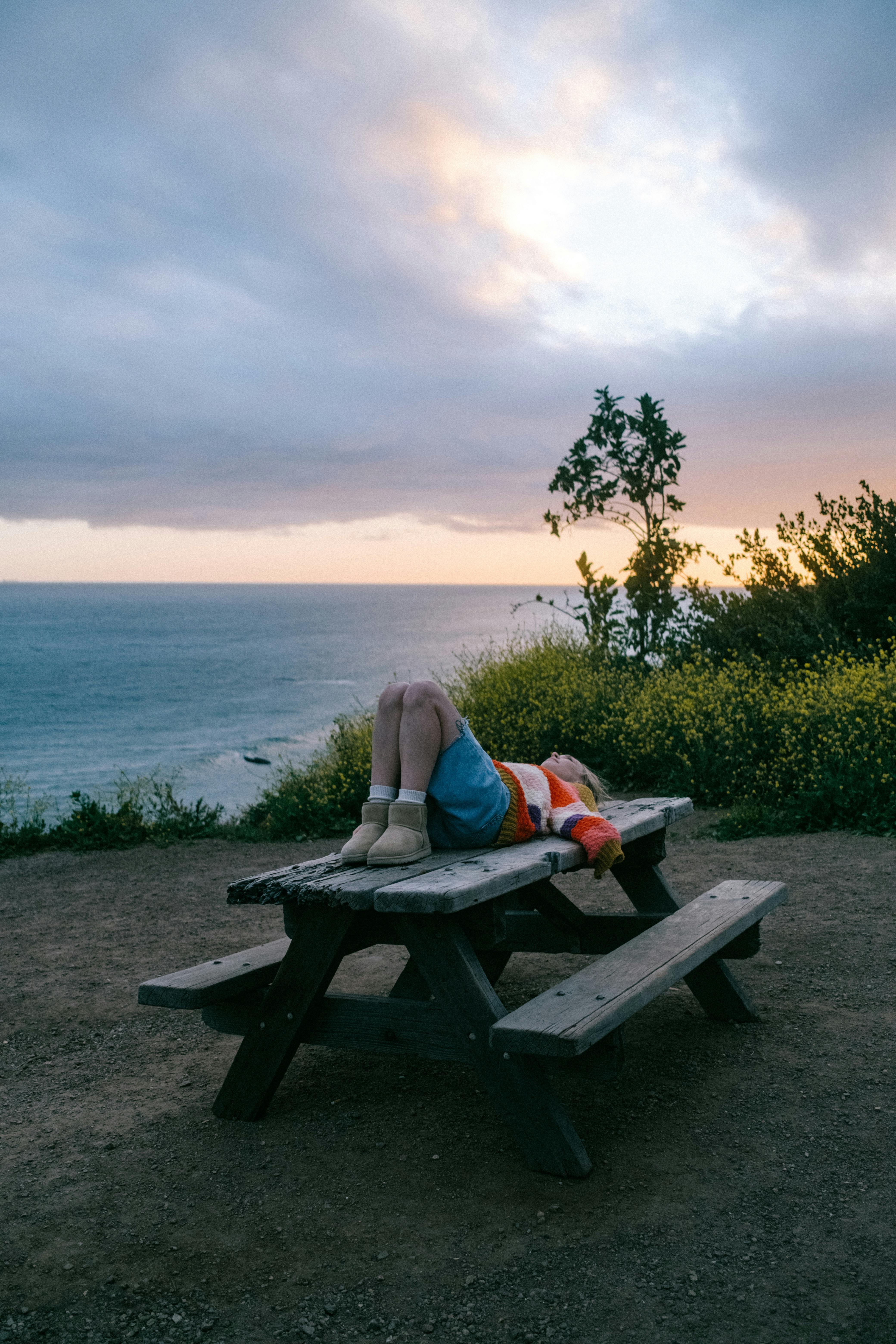 A person relaxing on a picnic table at sunset by the seashore. Perfect for themes of reflection and tranquility.