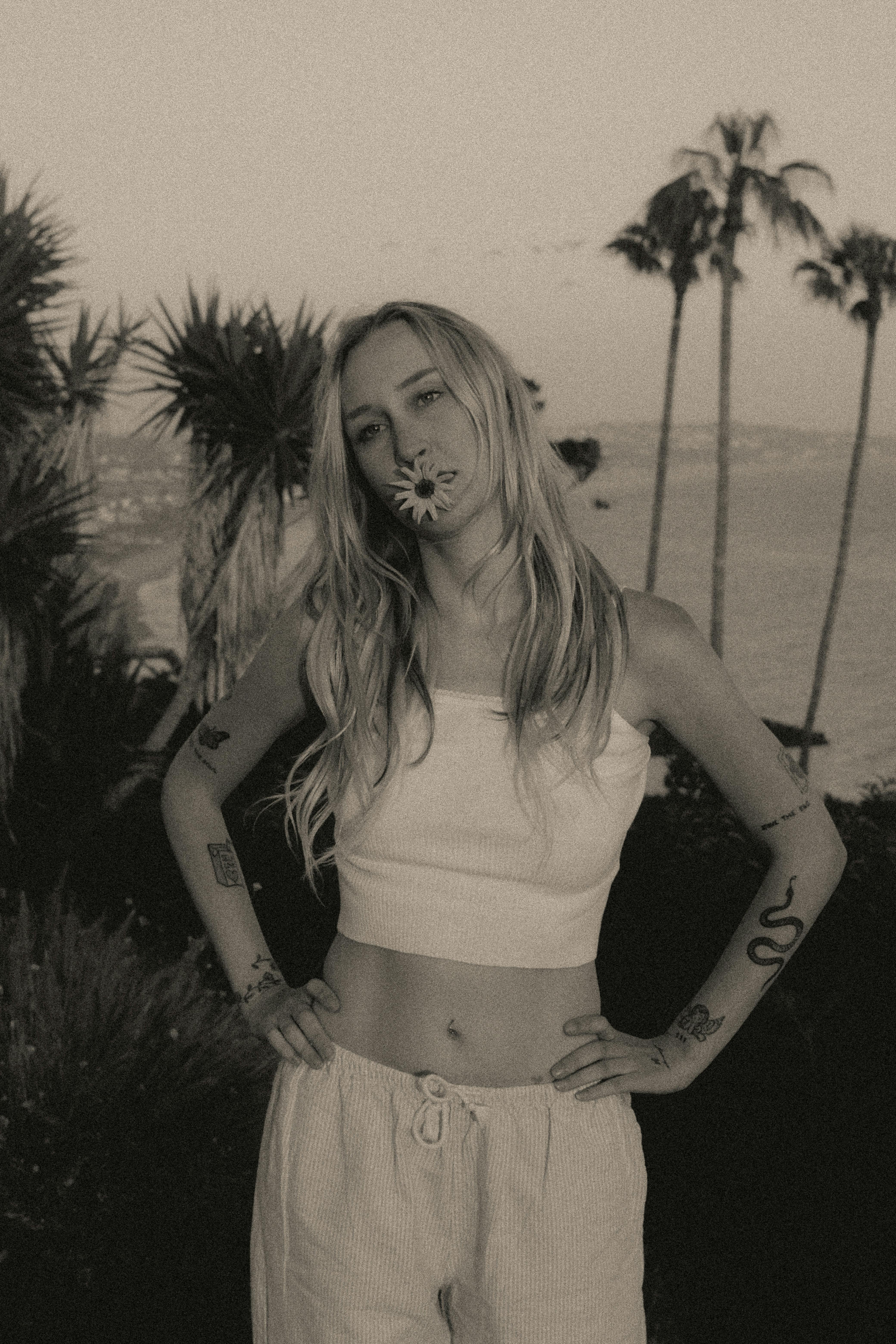 Woman with tattoos holding a flower in her mouth by the ocean.