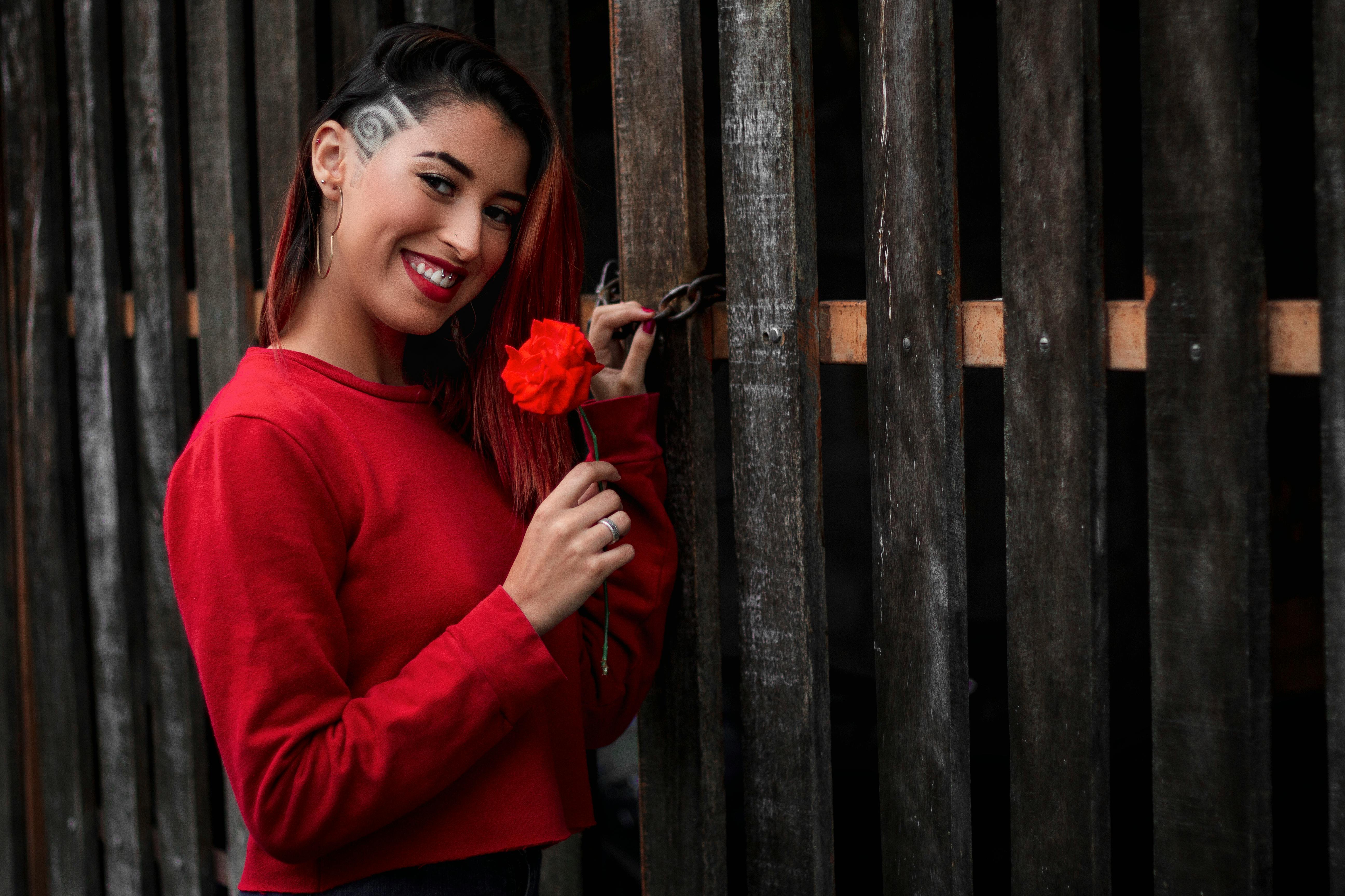 一位身穿红色毛衣的年轻女子手持玫瑰，在木栅栏旁微笑