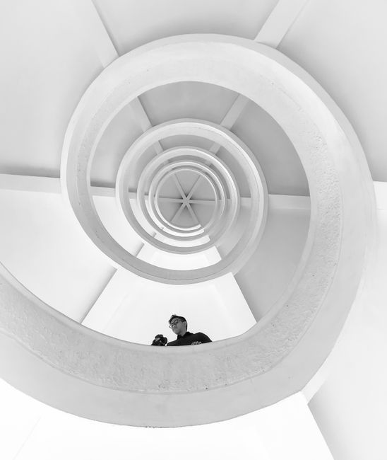 Monochrome image of a spiral staircase with a person looking up, showcasing geometric architecture.