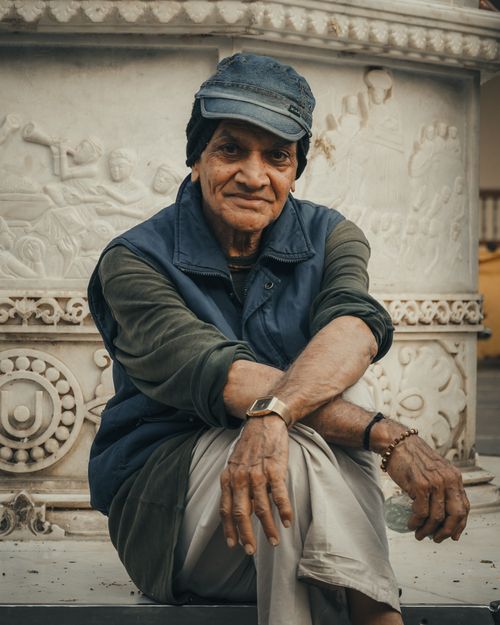Free Portrait of an elderly man in Ahmedabad, India, wearing a cap and vest, sitting against ornate stonework. Stock Photo