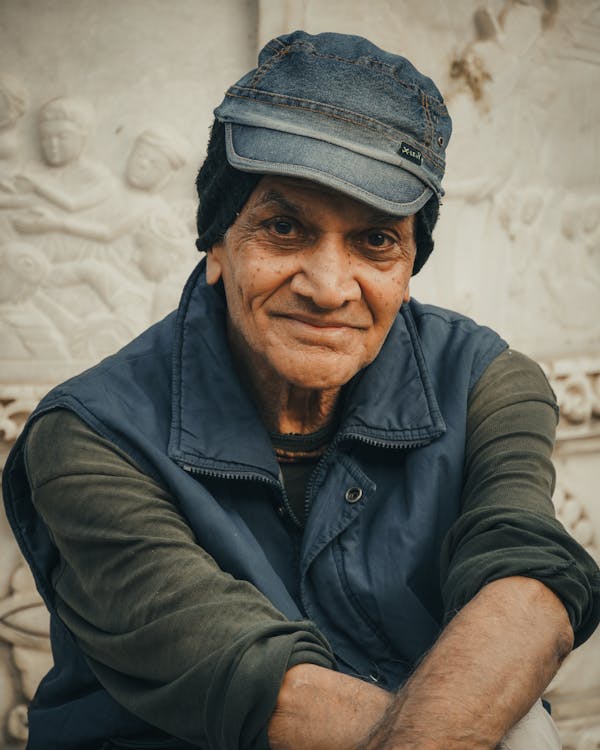 An elderly man sits on a stone bench in front of intricate temple pillars, smiling mysteriously.