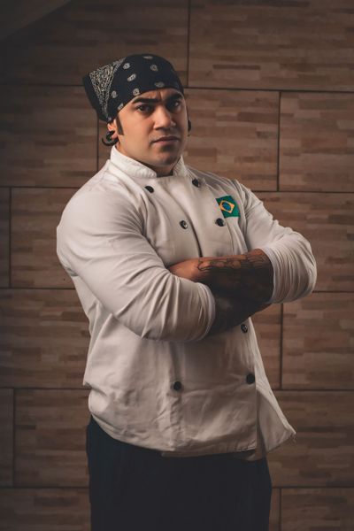 Portrait of a chef in uniform, standing confidently with arms crossed, wearing a bandana, set against a wooden background.