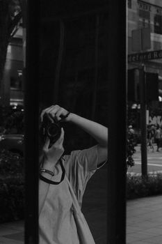 A black and white photo of a person taking their own picture in a reflective surface outdoors.