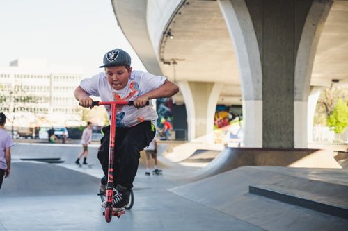 Free A young boy performing tricks on his kick scooter at an urban skatepark during the day. Stock Photo