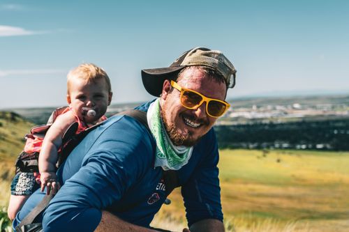Free Smiling dad with sunglasses hiking outdoors carrying a baby on his back. Stock Photo