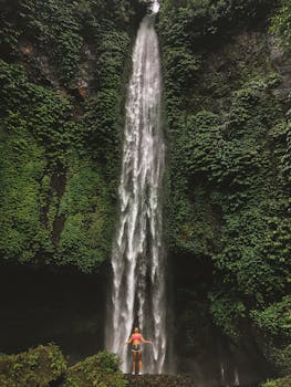 在巴厘岛雨林郁郁葱葱的植被环绕中，一个人孤零零地站在高耸的瀑布前