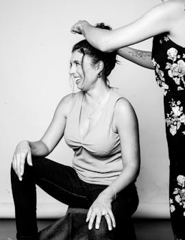 A woman joyfully laughs while getting her hair styled in an updo, captured in a black and white photo.