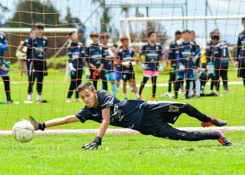 A young goalkeeper dives to save a soccer ball during a practice match outdoors.
