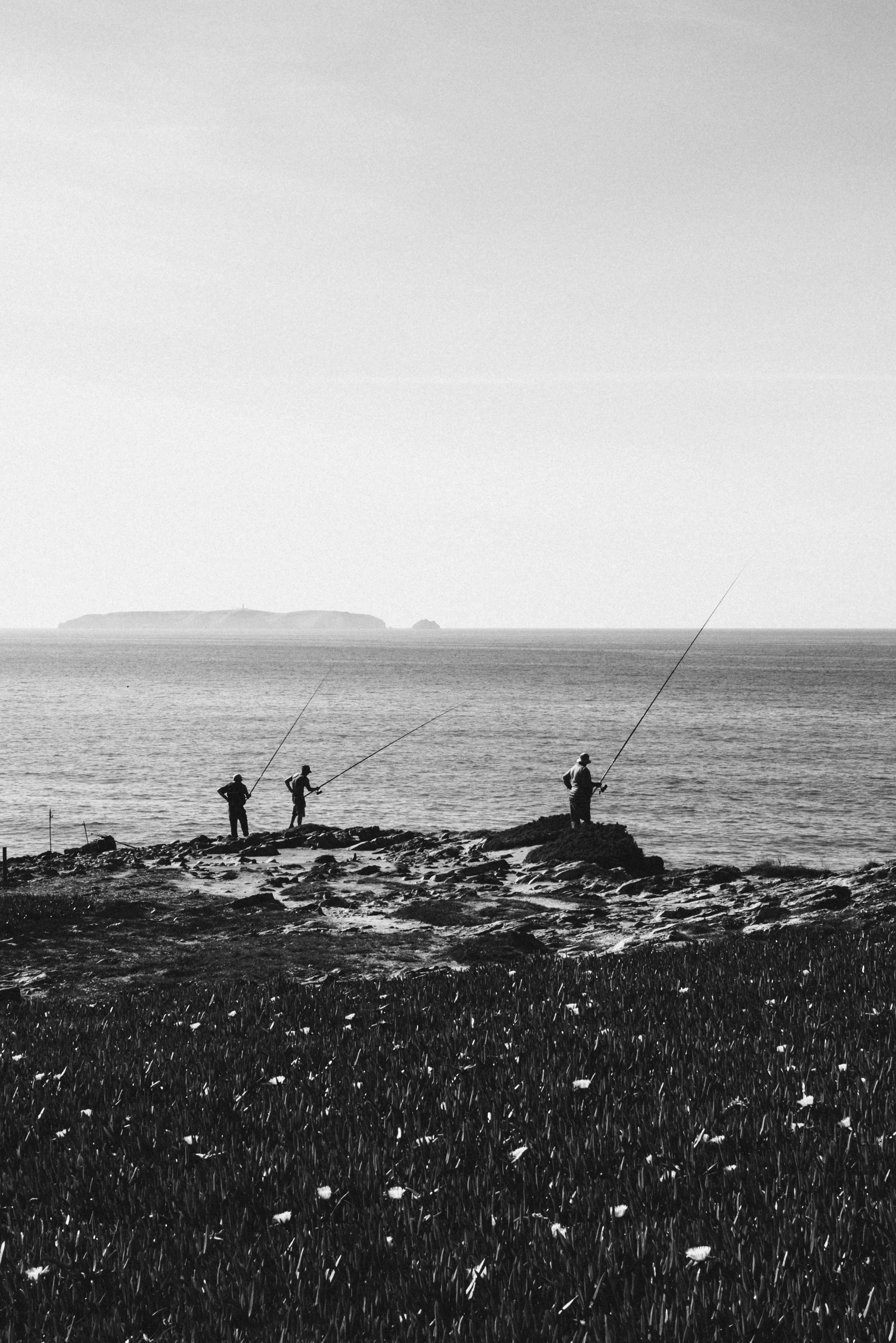 Three people fishing by the coast in black and white, showcasing a serene hobby.