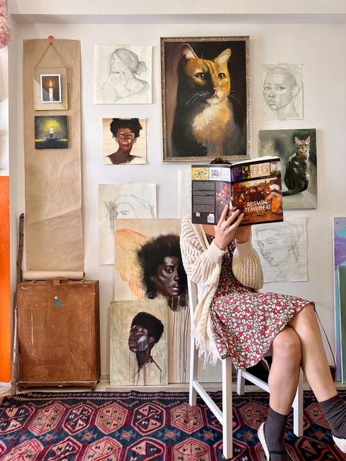 A woman in a floral dress reads a book in an art studio surrounded by various paintings and sketches.