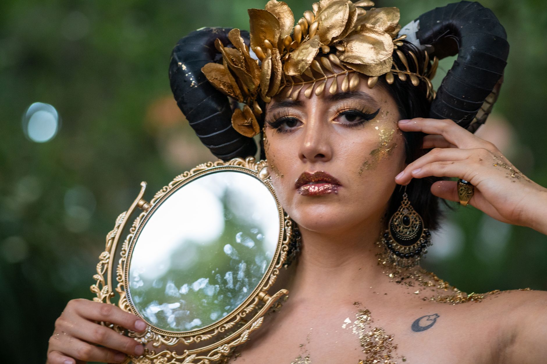 Portrait of a mythical woman adorned with golden horns, holding an ornate mirror outdoors.