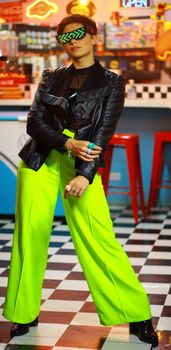 Young woman in a black jacket and neon pants posing in a retro diner with a futuristic twist.
