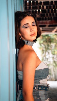 Stylish young woman with makeup and denim outfit posing against a vibrant wall in a sunlit space.