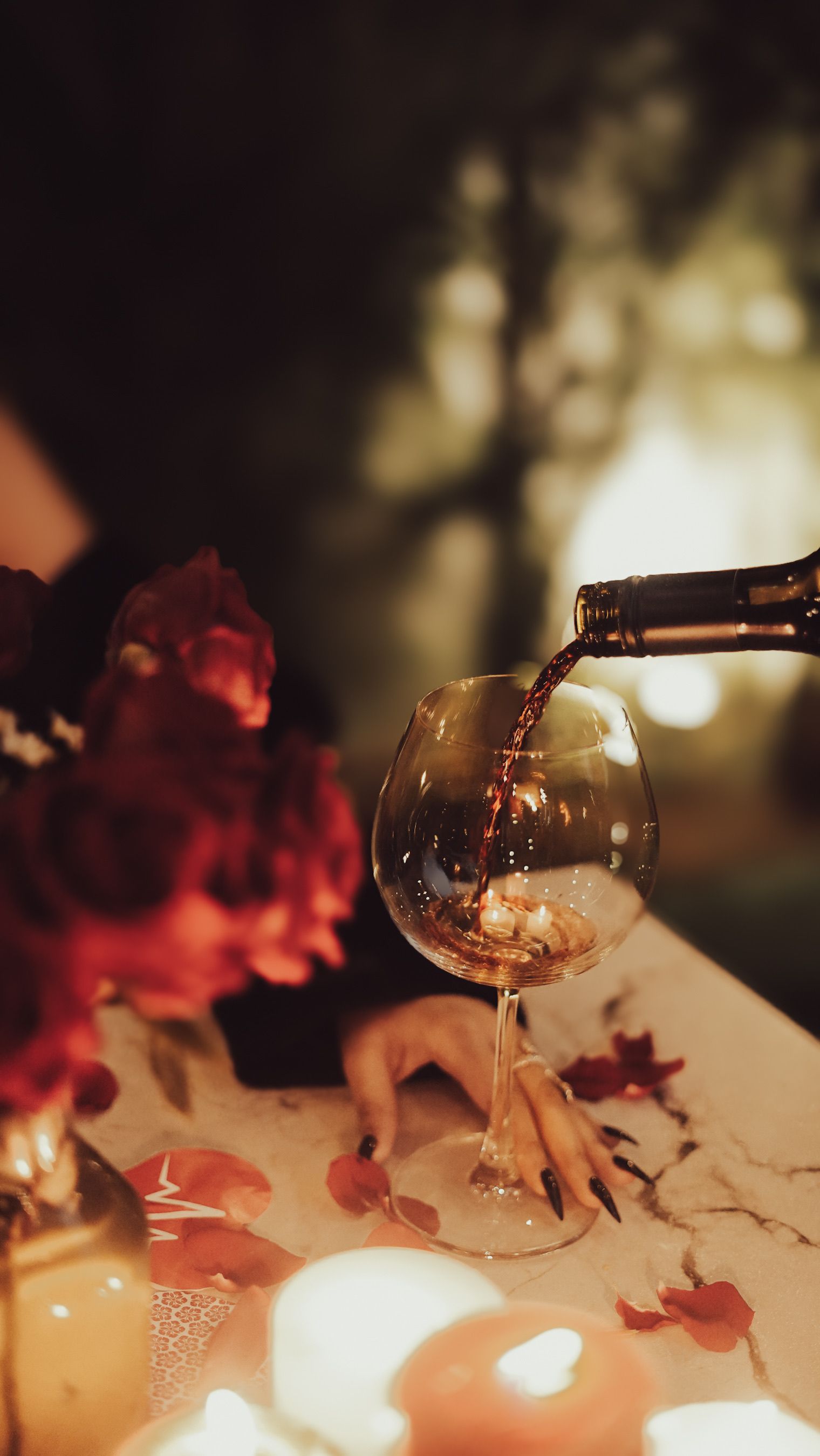 A cozy romantic setting with wine being poured into a glass amidst roses and candles.